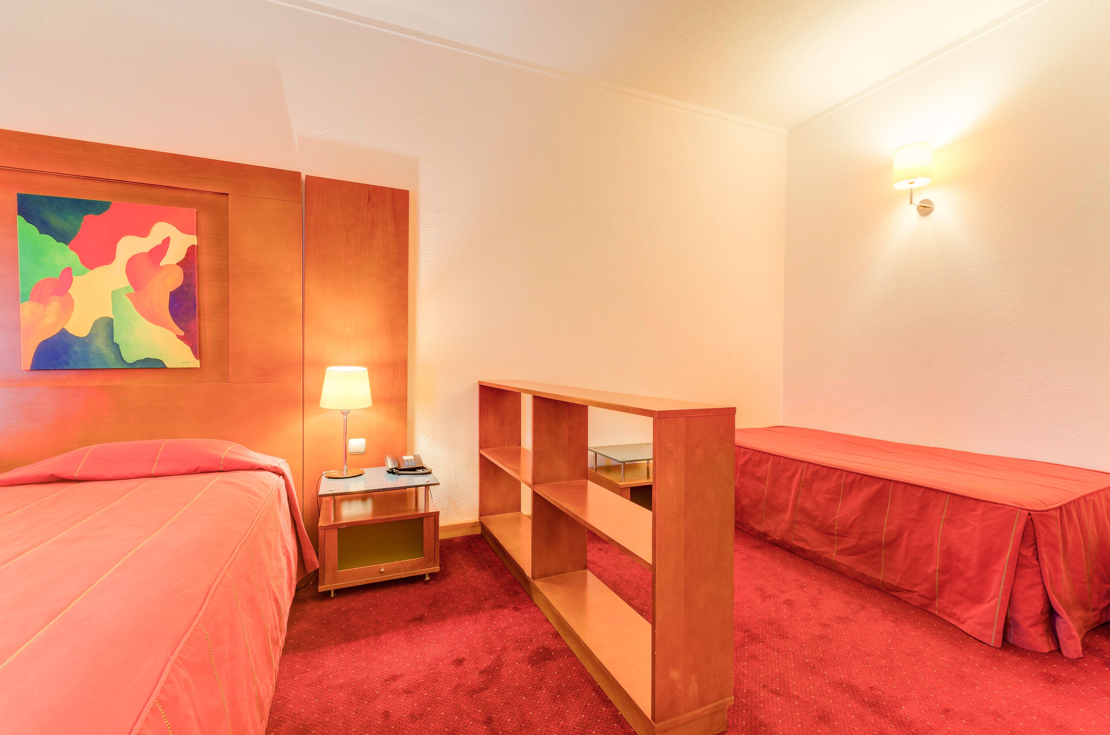 A hotel room with a bed, a desk, a lamp, a shelf, and a colorful abstract painting.