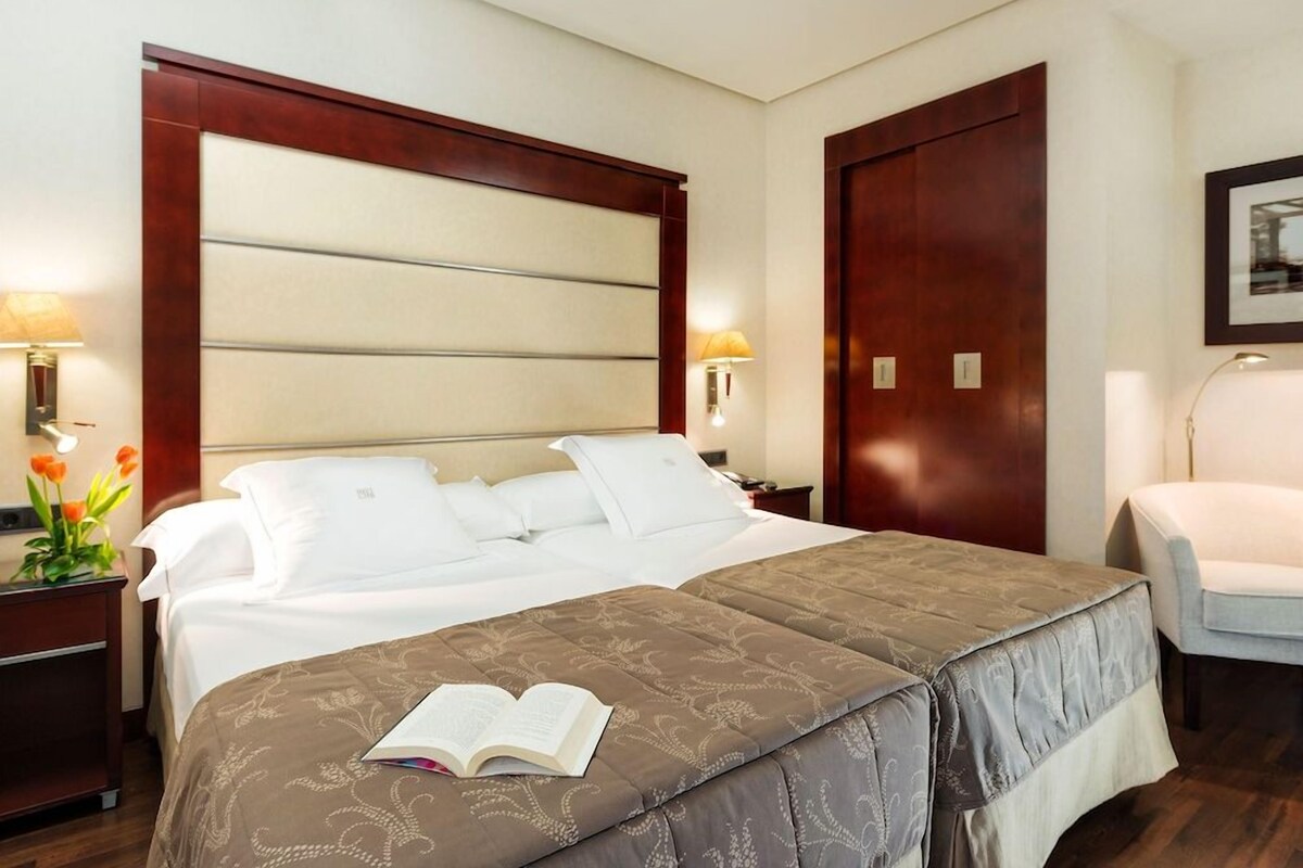 A hotel room with a large bed, a bedside table with a lamp, a chair, and a framed picture on the wall.