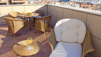 A rooftop terrace with wicker furniture, a table with fruit and drinks, and a city view.
