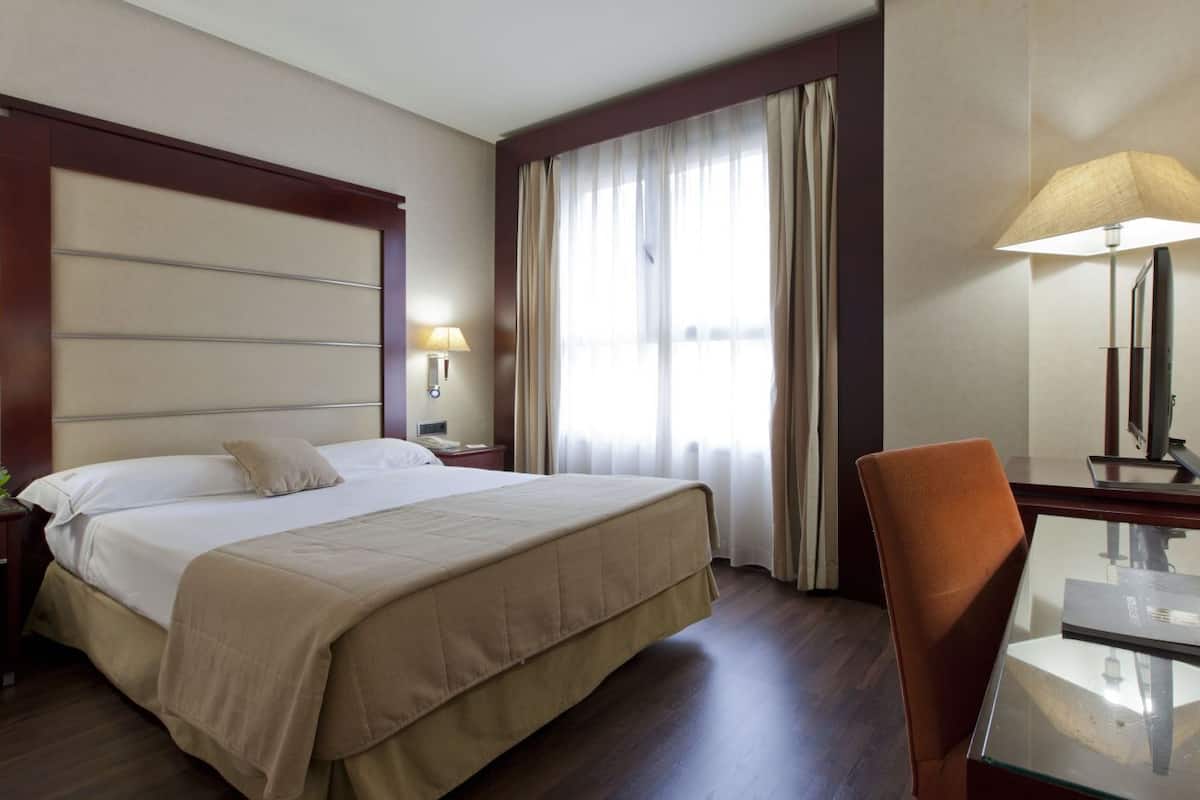 A hotel room with a large bed, bedside tables, a desk with a chair, and a window with curtains.