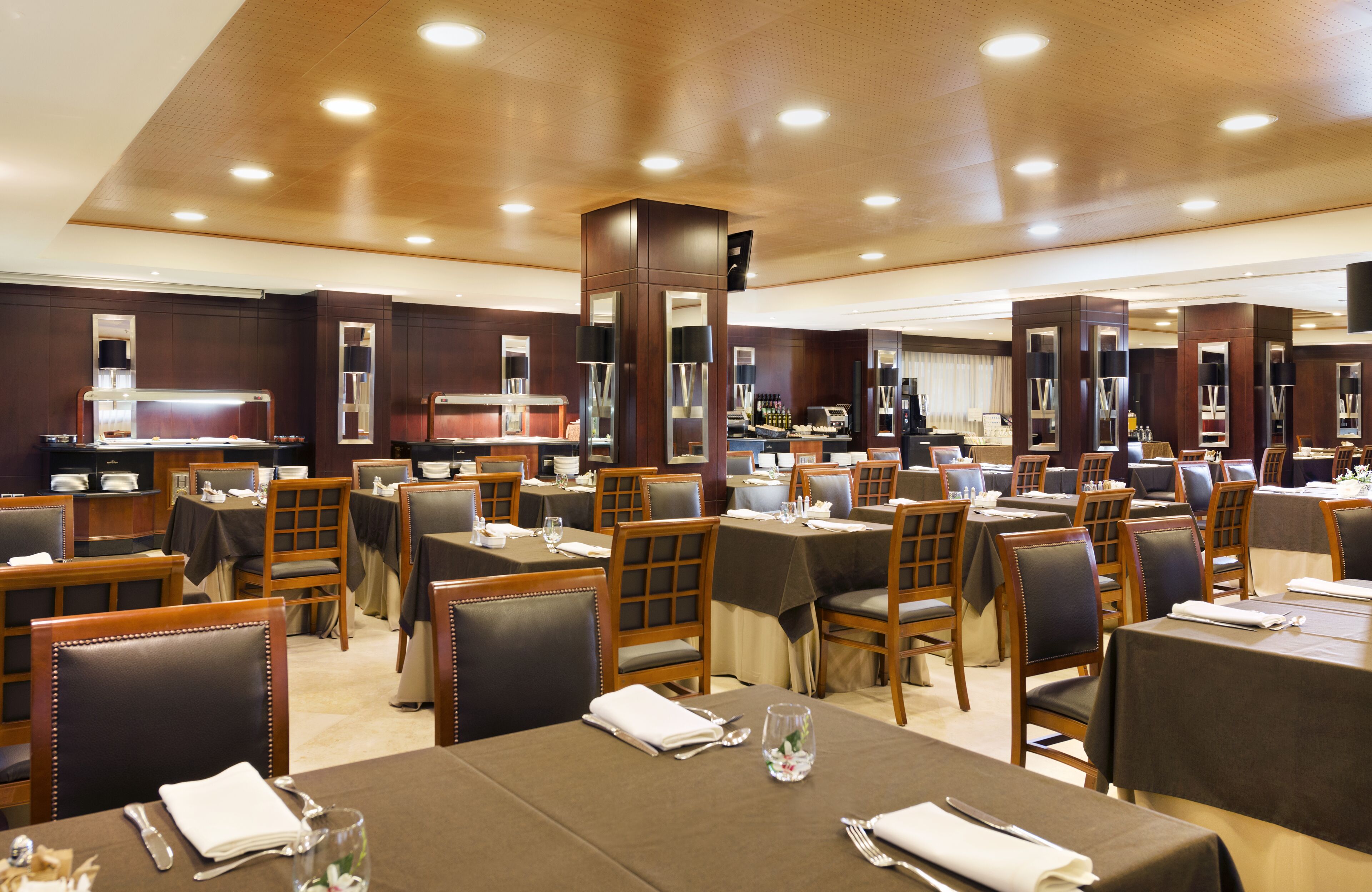 A spacious restaurant with multiple dining tables and chairs, a central bar area, and a modern ceiling design.