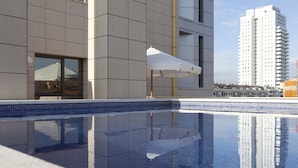 A modern building with a reflective pool, a white umbrella, and a tall adjacent building.