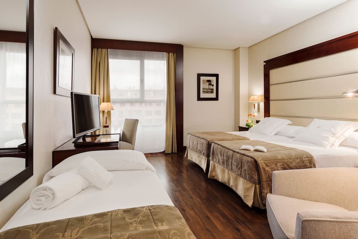 Ein Hotelzimmer mit zwei Betten, einem Schreibtisch mit Fernseher, einem Sessel und einem großen Fenster mit Vorhängen.