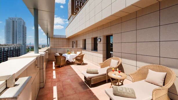 A rooftop terrace with wicker furniture, a table set with fruits and glasses, and a view of the cityscape.