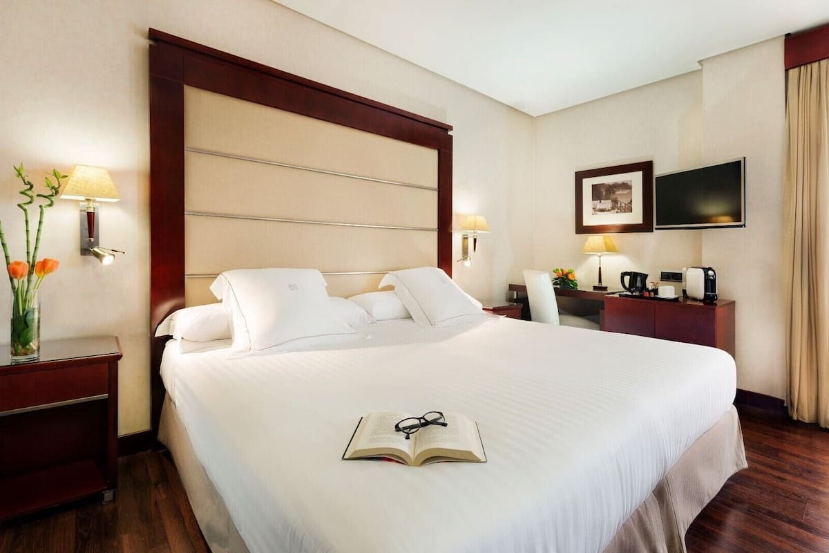 Ein Hotelzimmer mit einem großen Bett, Nachttischen, einem Schreibtisch, einem Sessel, einem Fernseher und einem Fenster mit Vorhängen.
