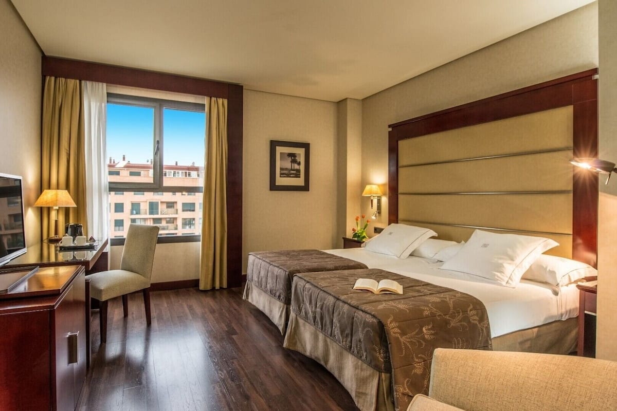 Ein Hotelzimmer mit Bett, Nachttisch, Lampe, Sessel, Fernseher und Blick auf Gebäude durch das Fenster.