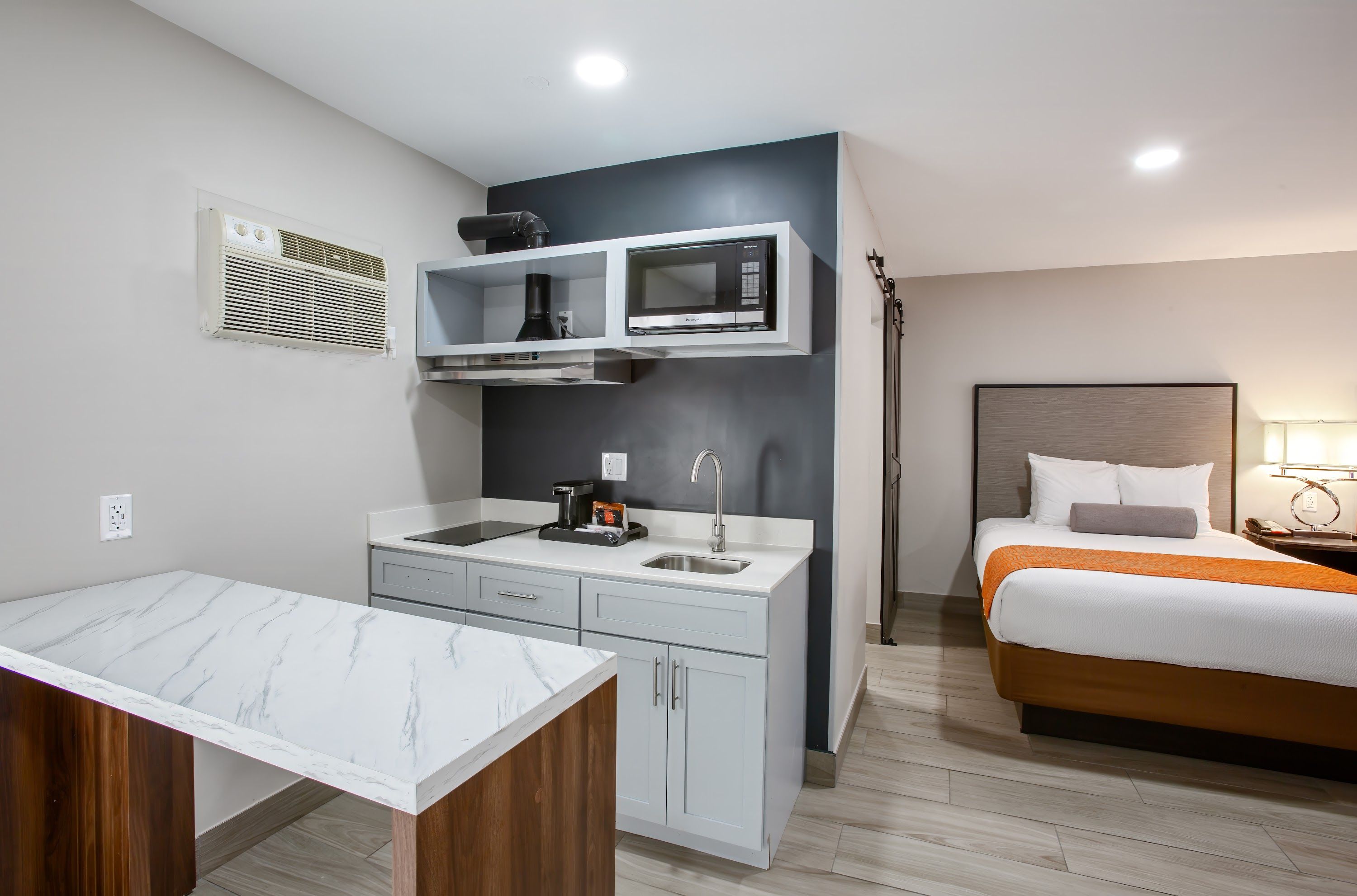 A compact hotel room with a kitchenette, a bed with an orange throw, and a marble-topped table.