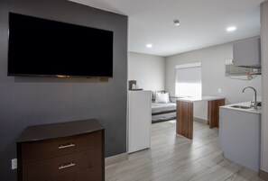 Habitación de hotel moderna y compacta con televisor de pantalla plana, cómoda de madera, kitchenette y cama con cabecera.