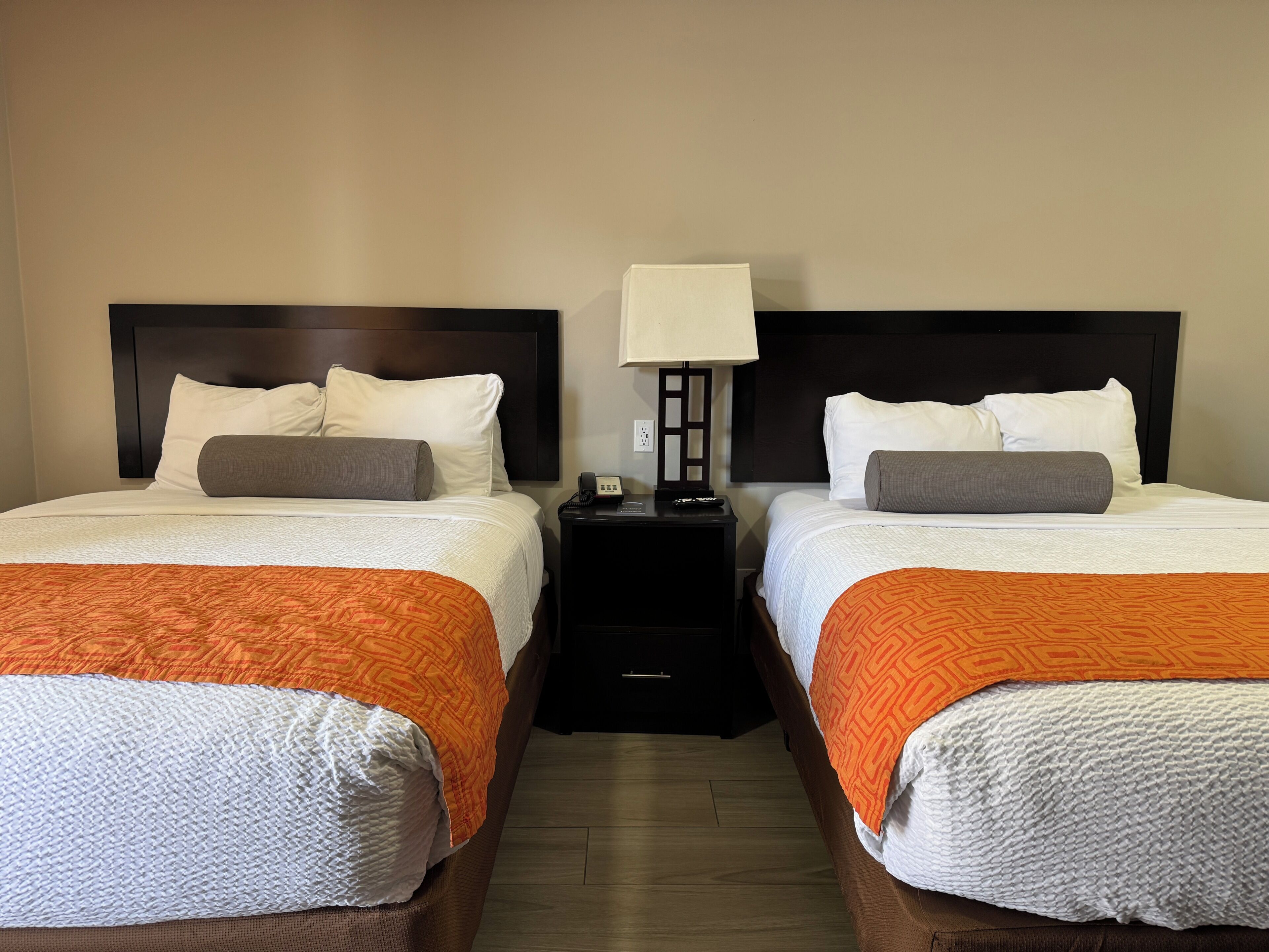 Une chambre d’hôtel avec deux lits, chacun recouvert d’une couette blanche, d’un plaid orange et doté d’une tête de lit en bois. Une table de chevet avec une lampe se trouve entre les deux lits.