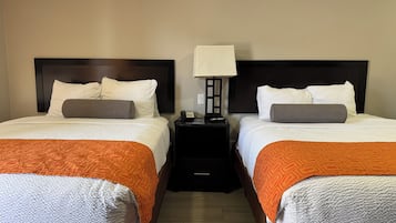 Habitación de hotel con dos camas, cada una con cubrecama blanco, manta naranja y cabecero de madera. Entre las camas, una mesita de noche con una lámpara.