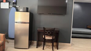A compact room with a refrigerator, a chair, a small table, and a flat-screen TV mounted on the wall.