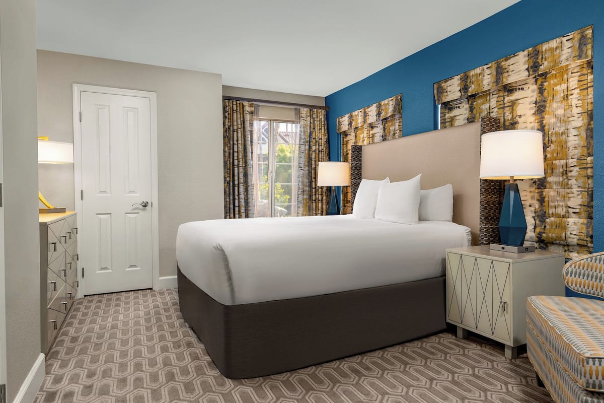 A hotel room with a large bed, two bedside tables with lamps, a chair, and a view of the outdoors through the window.