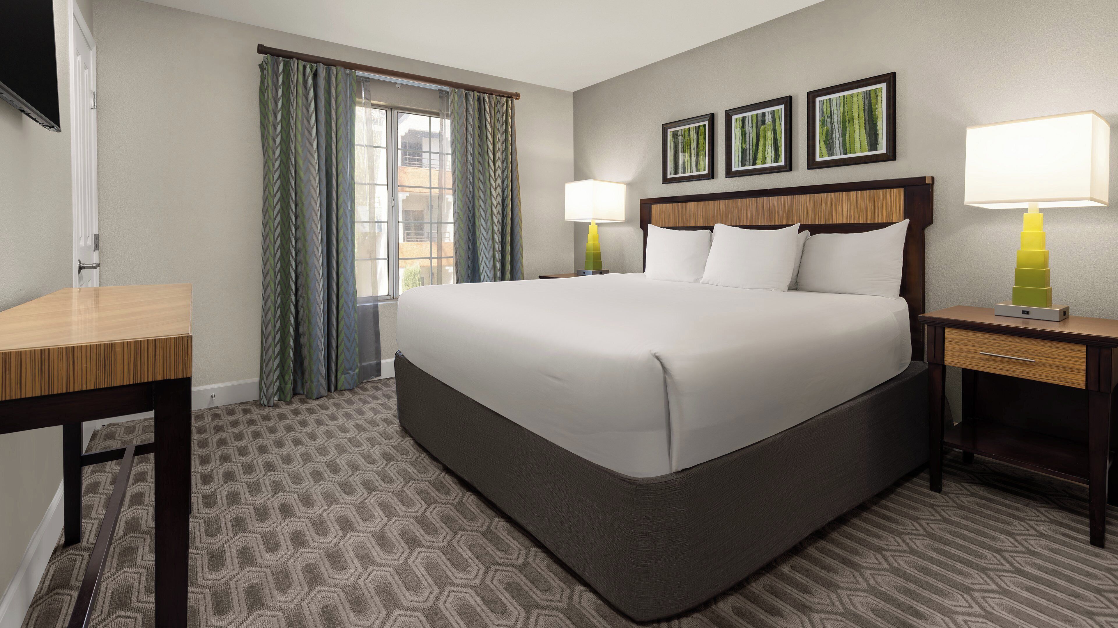 A hotel room with a large bed, a desk, a chair, a nightstand, and a window with curtains.