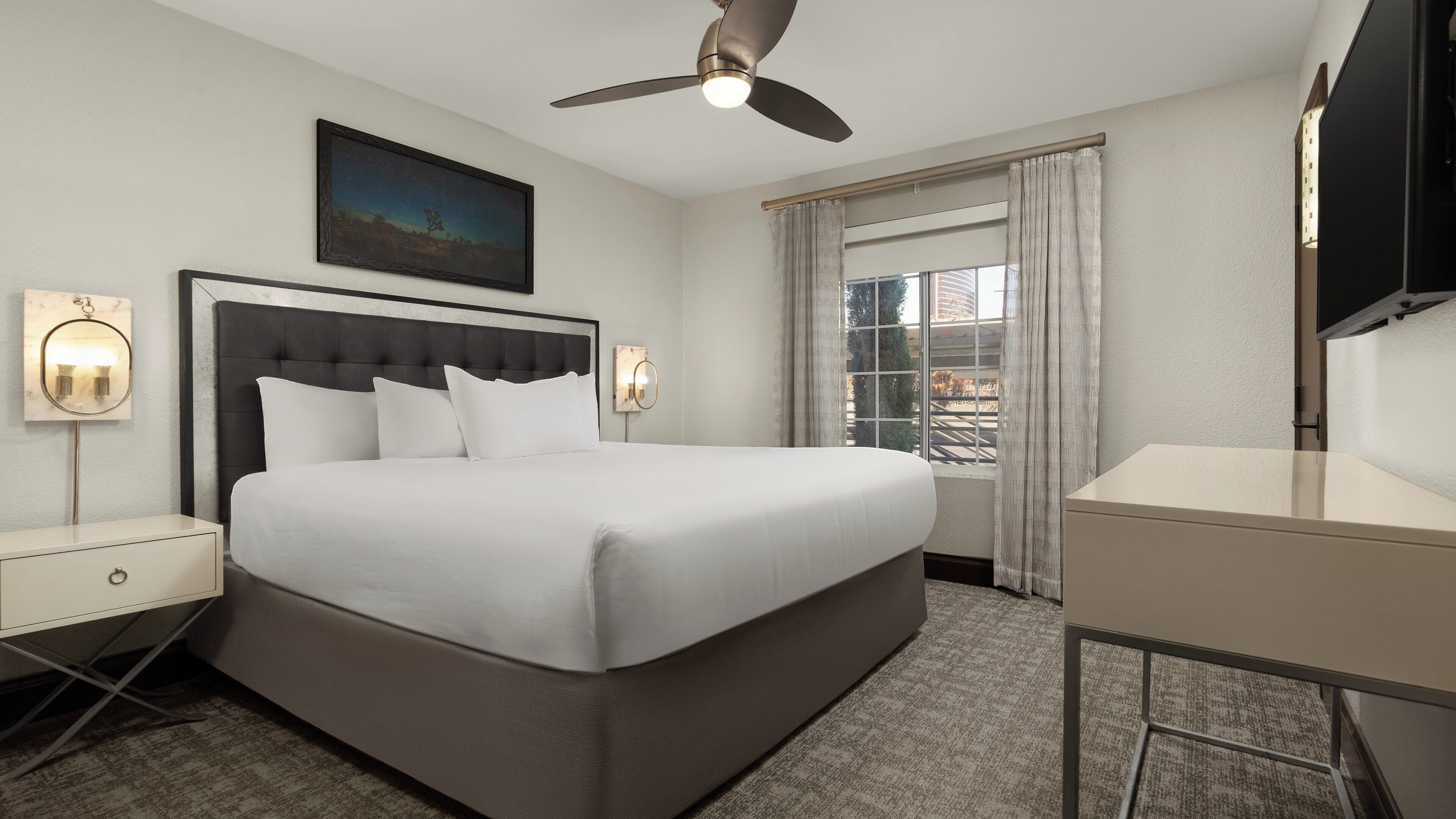 A hotel room with a large bed, a ceiling fan, a nightstand with a lamp, a desk, and a window with curtains.
