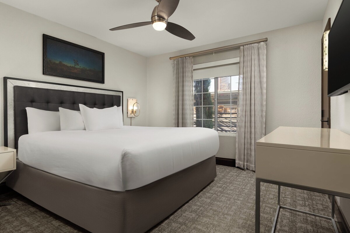 A hotel room with a large bed, a ceiling fan, a nightstand with a lamp, a desk, and a window with curtains.