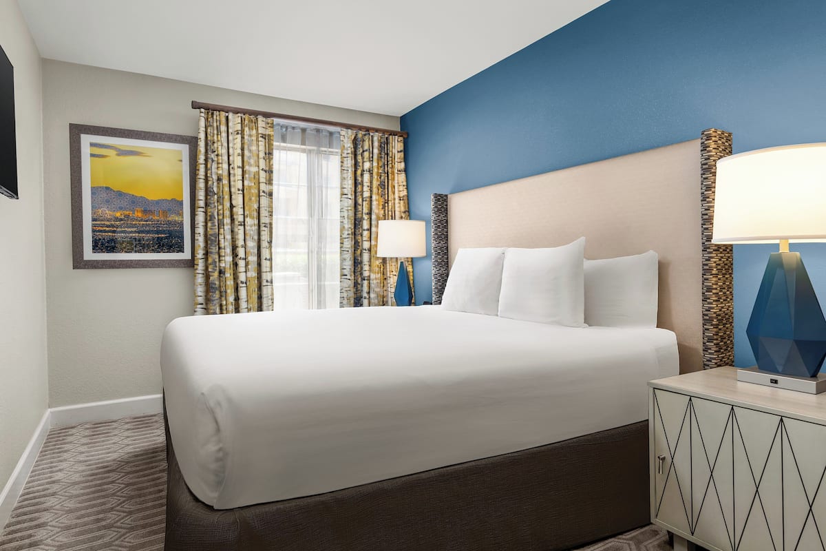 A hotel room with a large bed, a nightstand with a lamp, a wall-mounted TV, and a framed picture of a cityscape.