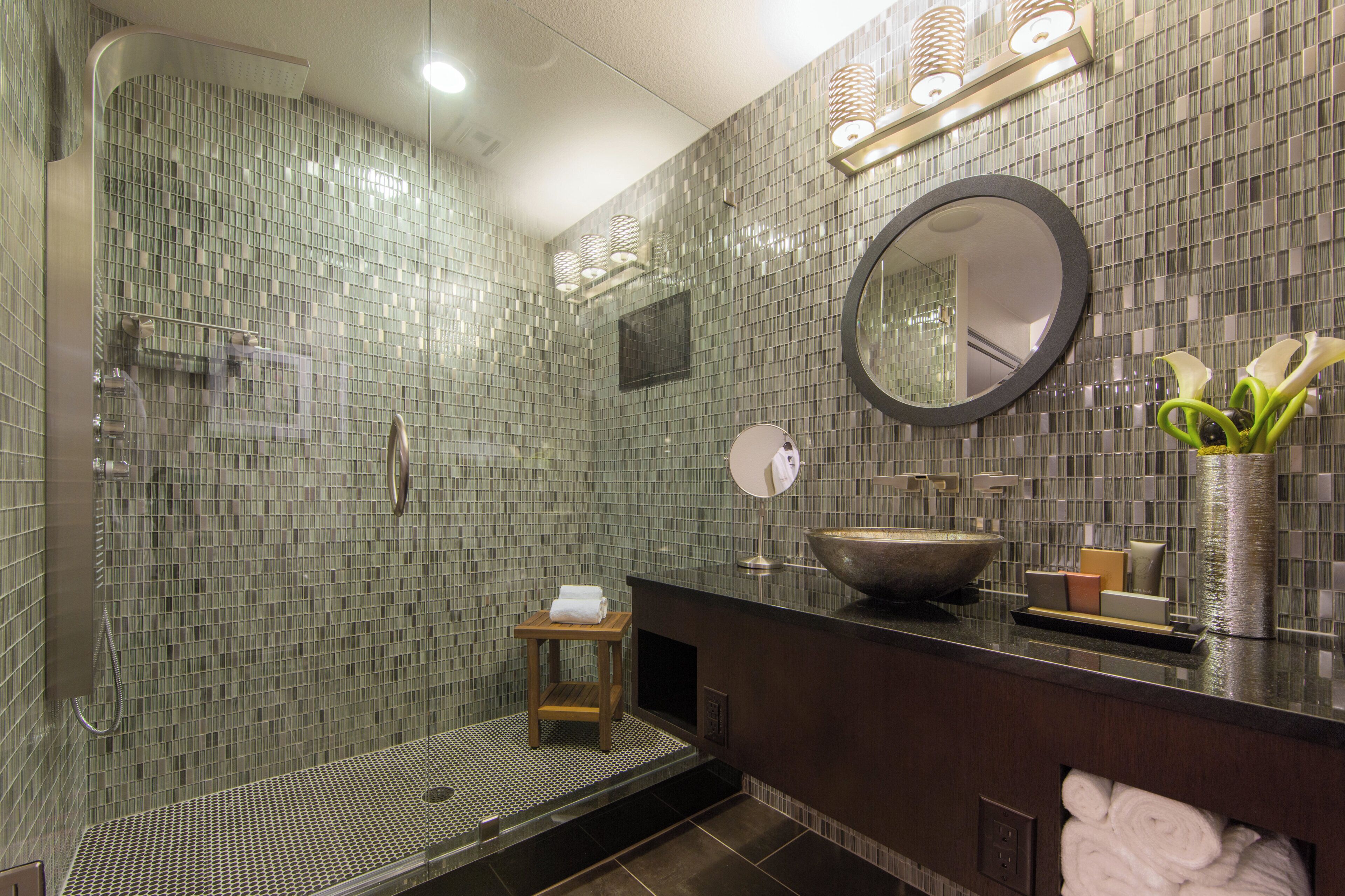 A modern bathroom with a glass shower enclosure, a round mirror, a large sink, and a small wooden stool.