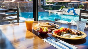 A dining table with breakfast items, a glass of orange juice, and a view of a swimming pool and umbrellas. - Holiday Inn Club Vacations at Desert Club Resort by IHG (Las Vegas)