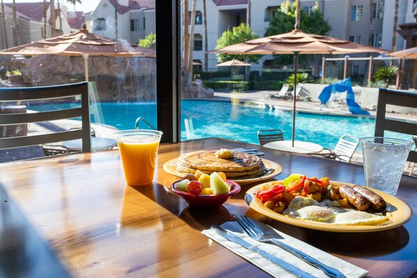 A dining table with breakfast items, a glass of orange juice, and a view of a swimming pool and umbrellas. - Holiday Inn Club Vacations at Desert Club Resort by IHG (Las Vegas)