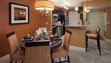 A dining area with a round table, chairs, and a chandelier. There is a kitchen in the background with a refrigerator and microwave.