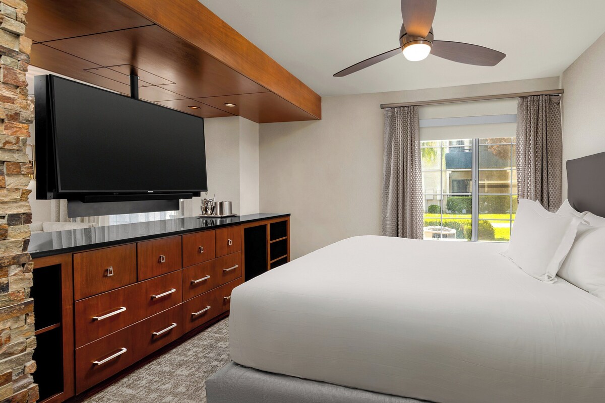 A modern hotel room with a large bed, wooden dresser, flat-screen TV, and a view of a garden through a window.