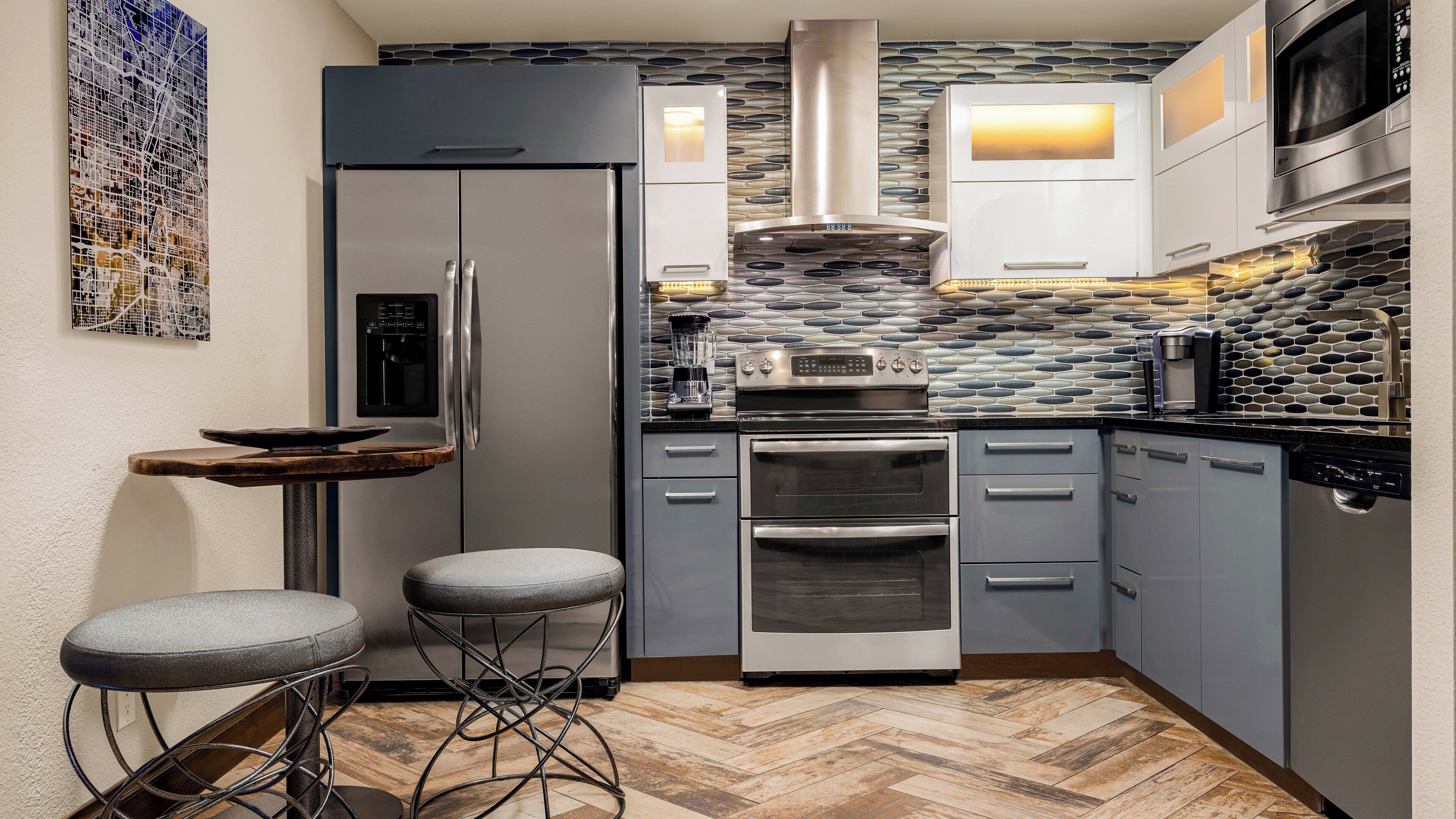 Eine kompakte Küche mit Kühlschrank, Backofen, Mikrowelle und einer Spritzschutzwand mit Steinmuster. Zwei Barhocker und ein kleiner Holztisch sind ebenfalls vorhanden.