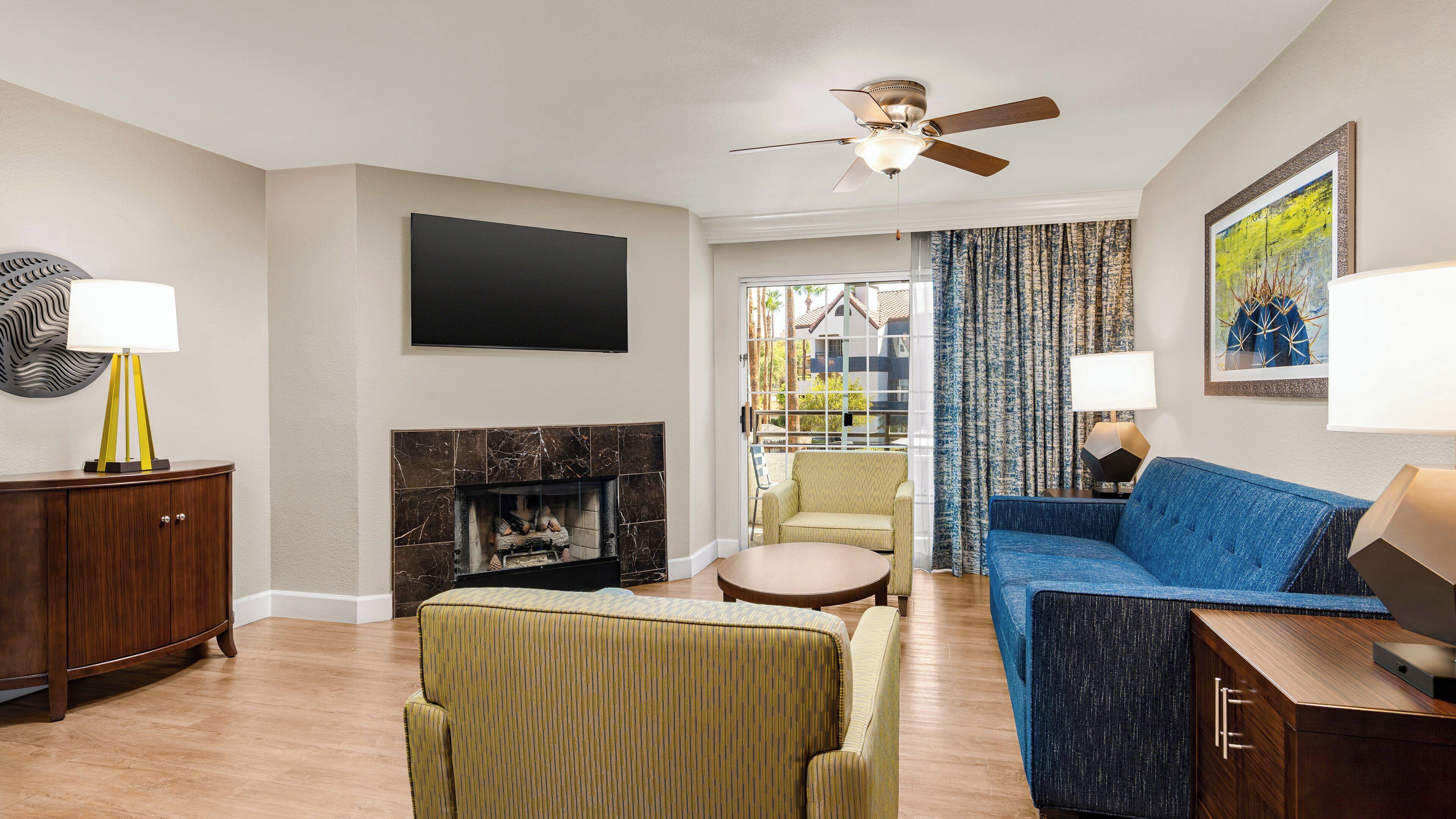 A living room with a fireplace, a blue sofa, a green armchair, a wooden cabinet, and a ceiling fan.