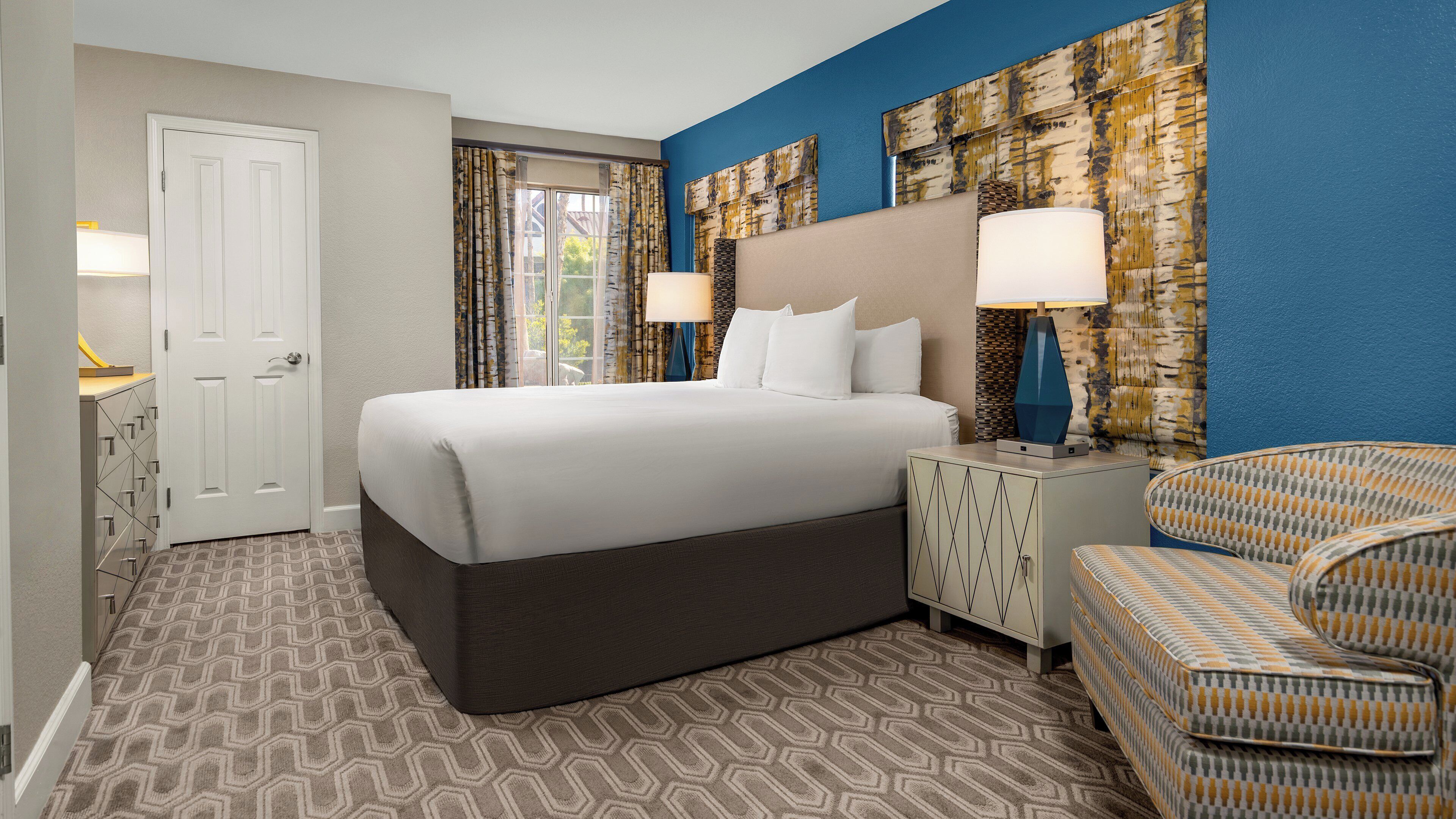 A hotel room with a large bed, two nightstands with lamps, a patterned armchair, and a view of the outdoors through the window.