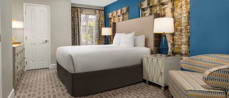A hotel room with a large bed, two nightstands with lamps, a patterned armchair, and a view of the outdoors through the window.