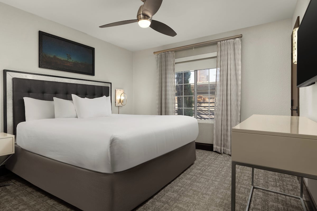 A hotel room with a large bed, a ceiling fan, a nightstand with a lamp, a desk, and a window with curtains.