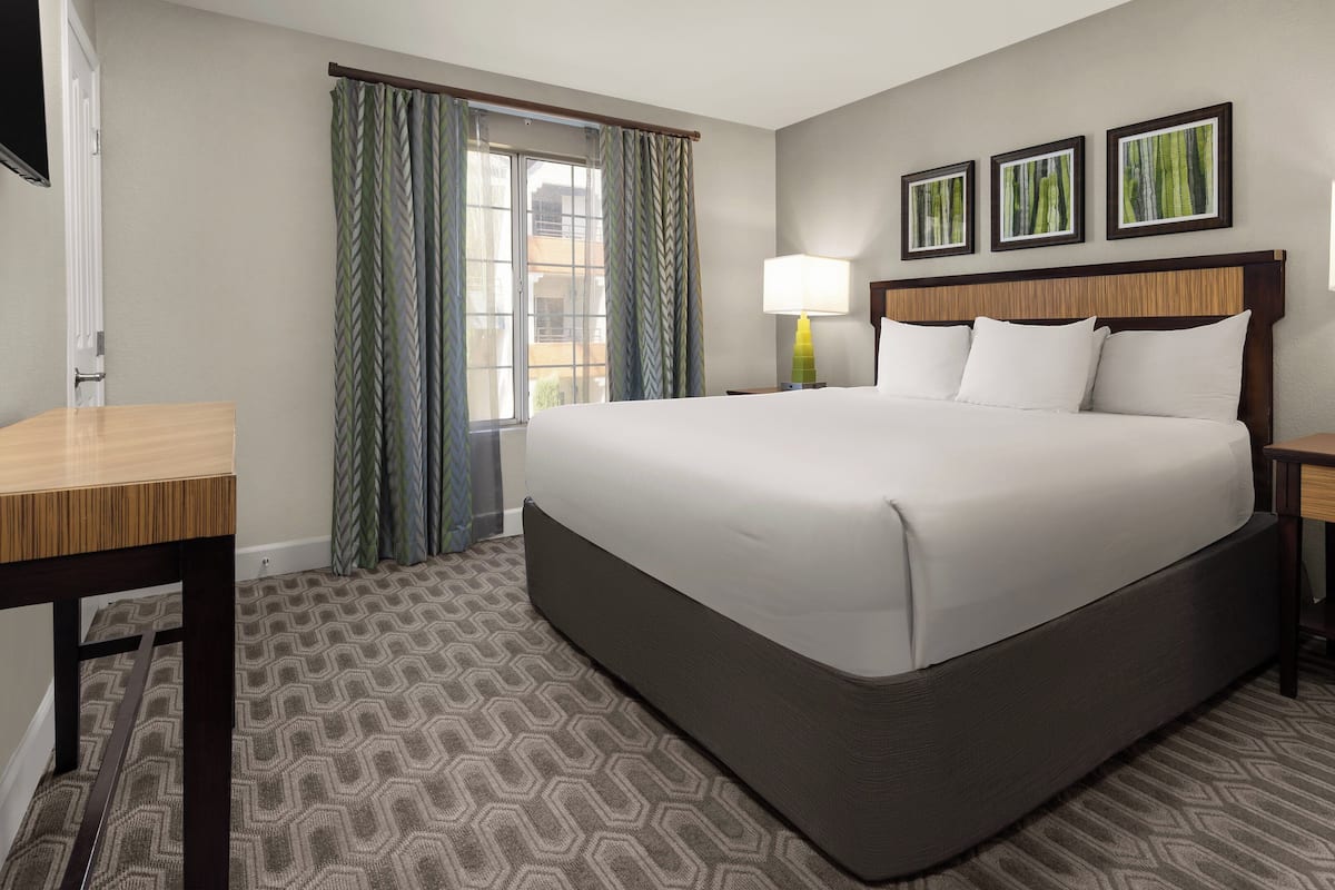 A hotel room with a large bed, a desk, a chair, a nightstand, and a window with curtains.