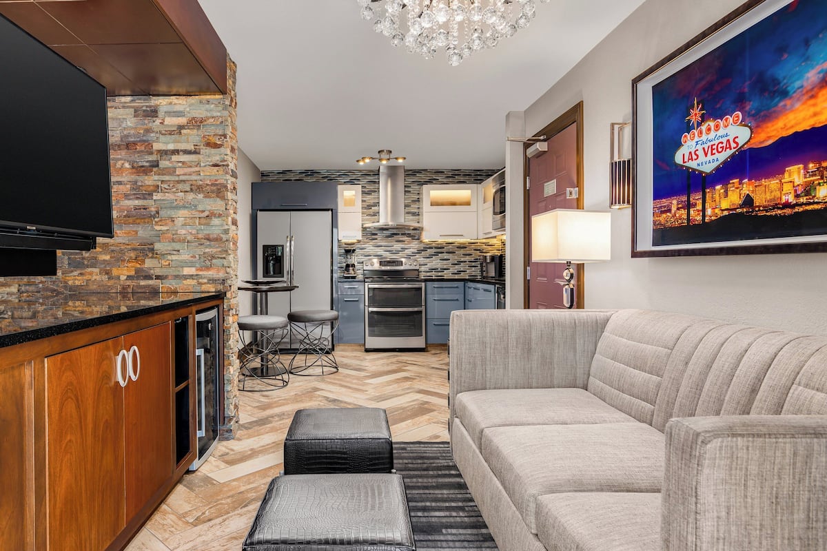 A modern living room with a flat-screen TV, a stone accent wall, a kitchen in the background, and a framed Las Vegas sign.