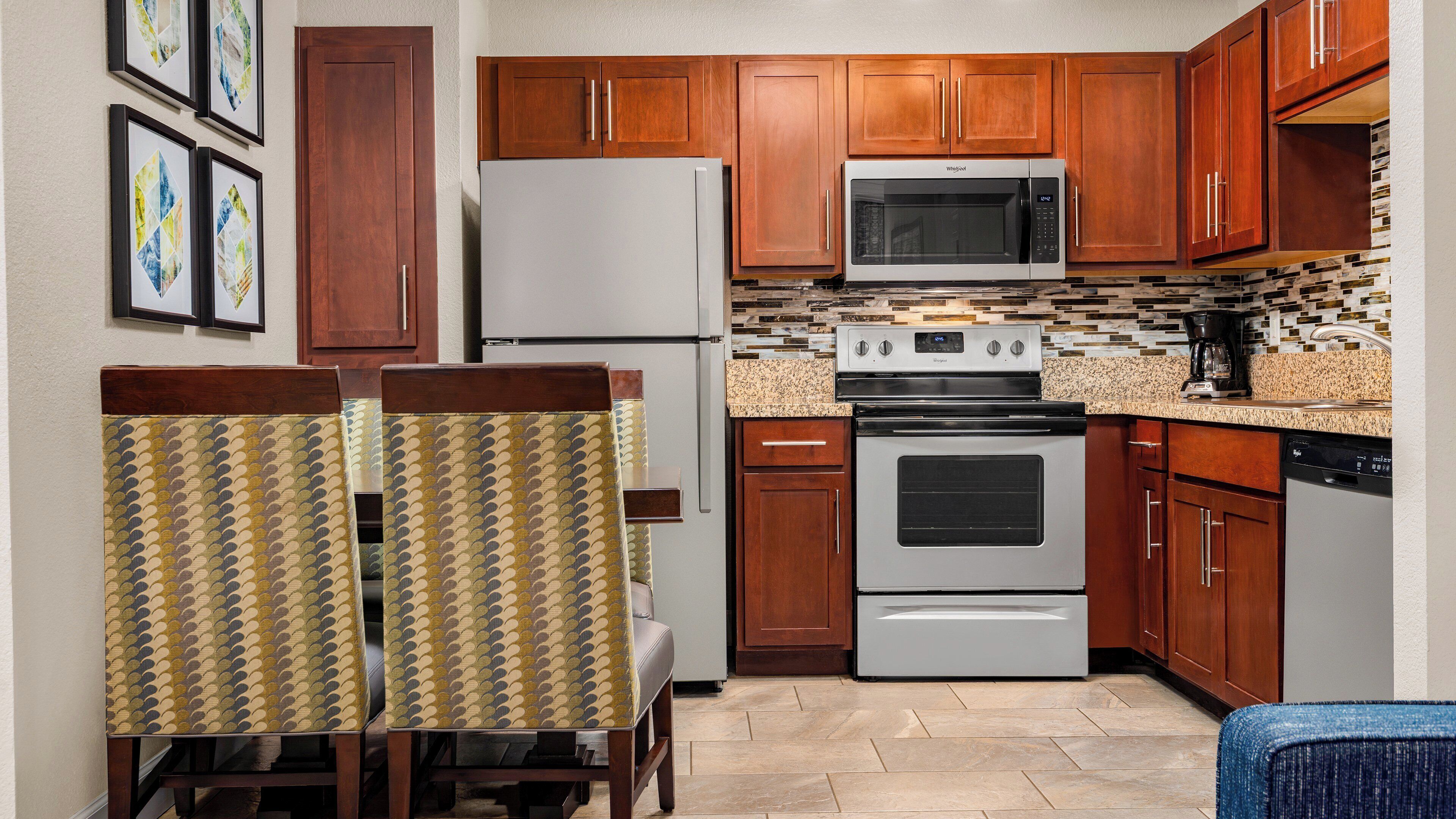 A compact kitchen with wooden cabinets, a built-in microwave, a stove, and a refrigerator. There are two chairs with patterned upholstery and a small table.