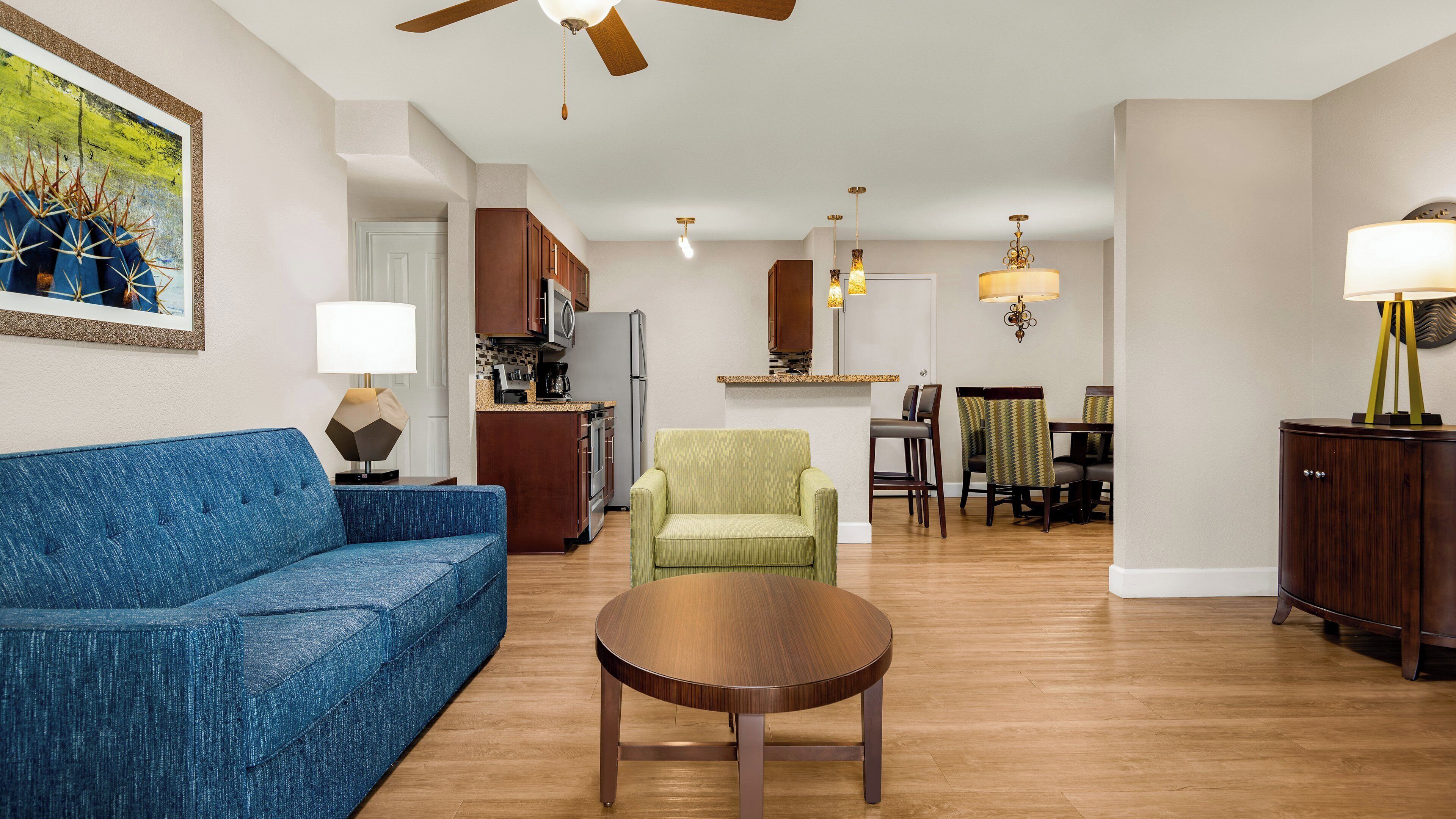 A living room with a blue sofa, a round coffee table, and a green armchair. There is a kitchen in the background with wooden cabinets and a dining area with a table and chairs.
