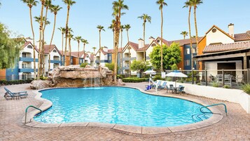 A swimming pool with a waterfall feature, surrounded by palm trees and a poolside area with lounge chairs and umbrellas.