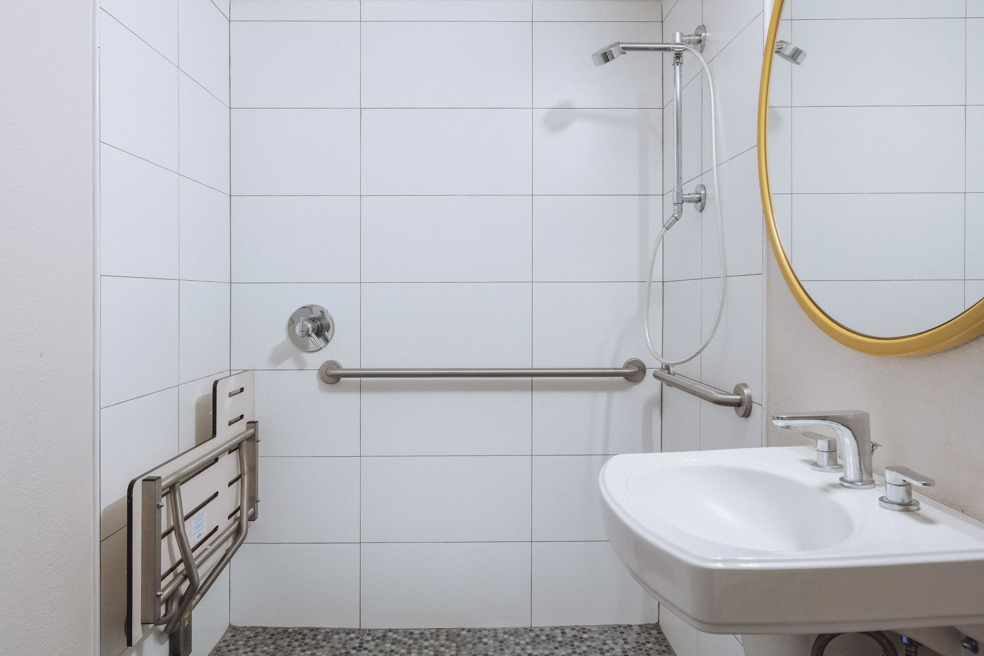 Uma casa de banho com paredes revestidas a azulejos brancos, uma pia branca, um espelho com moldura dourada e um chuveiro com barra de mão.