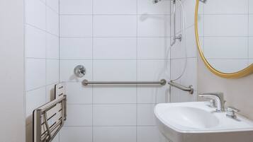Uma casa de banho com paredes revestidas a azulejos brancos, uma pia branca, um espelho com moldura dourada e um chuveiro com barra de mão.