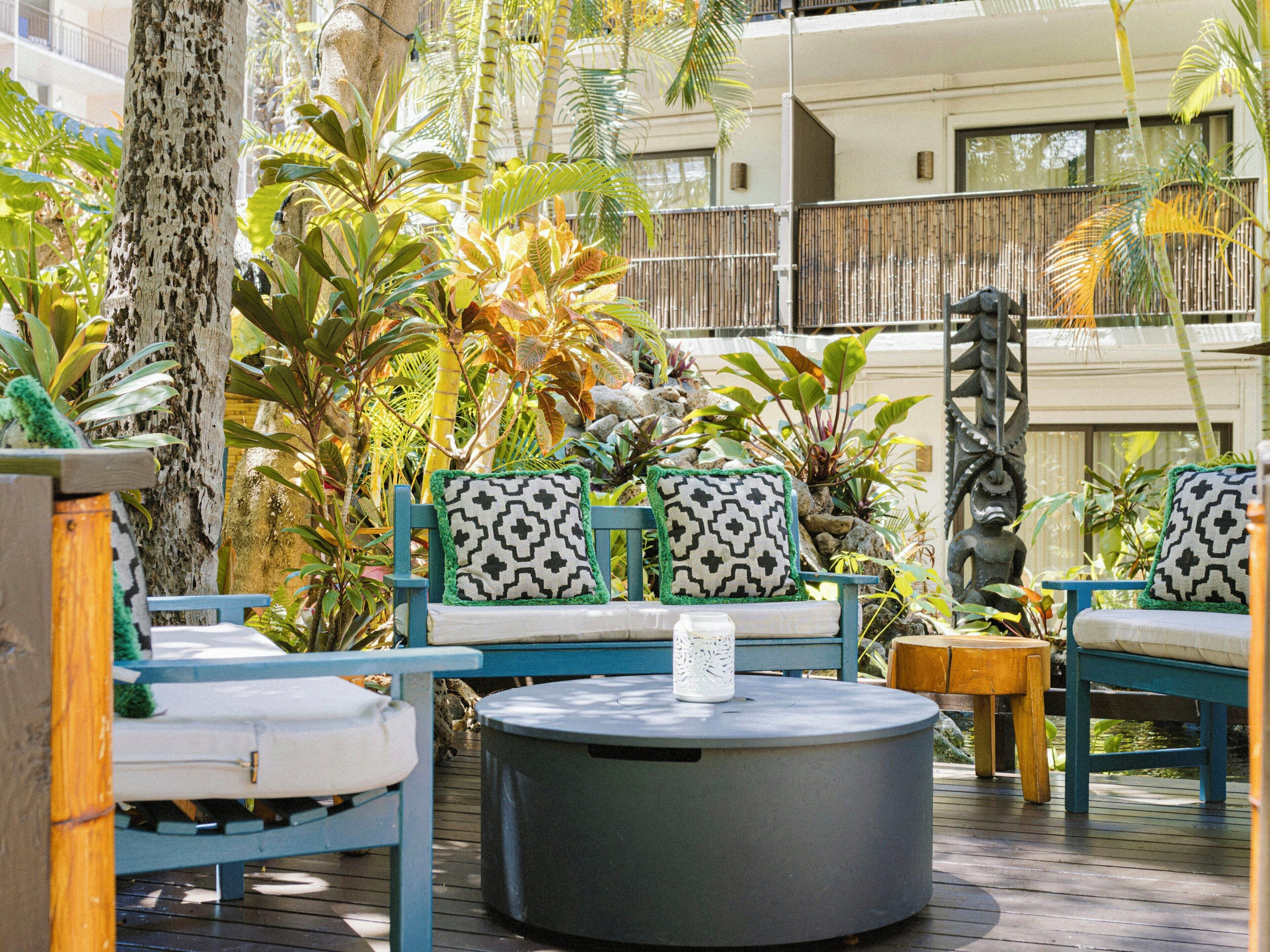 Un área de estar al aire libre con mesas y sillas redondas, rodeada de plantas tropicales y una terraza de madera.