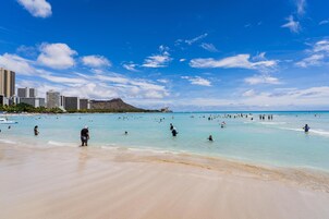Una playa con agua azul cristalina, gente nadando y caminando por la orilla, y edificios al fondo.