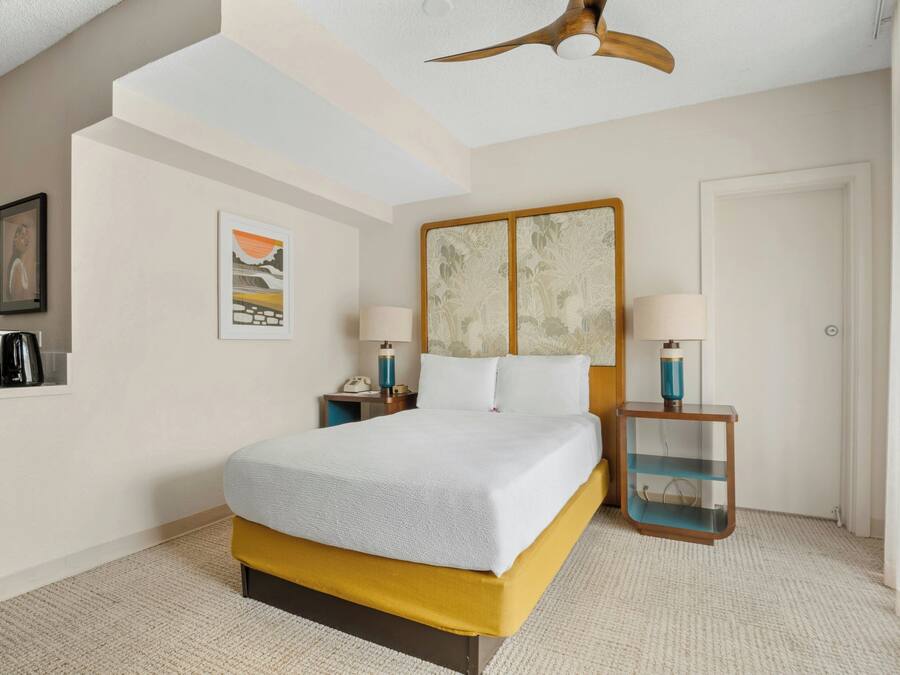 Una habitación de hotel con cama, mesitas de noche, ventilador de techo, un cuadro y una puerta.