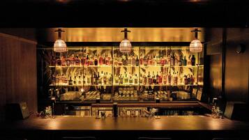 Zona de bar con estantes llenos de botellas y vasos, barra y lámparas colgantes.