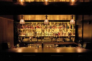 Zona de bar con estantes llenos de botellas y vasos, barra y lámparas colgantes.