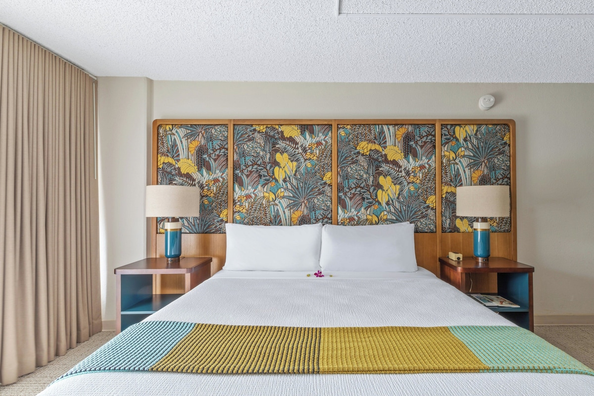 A hotel room with a large bed, two bedside tables, a headboard with a floral pattern, and a window with curtains.