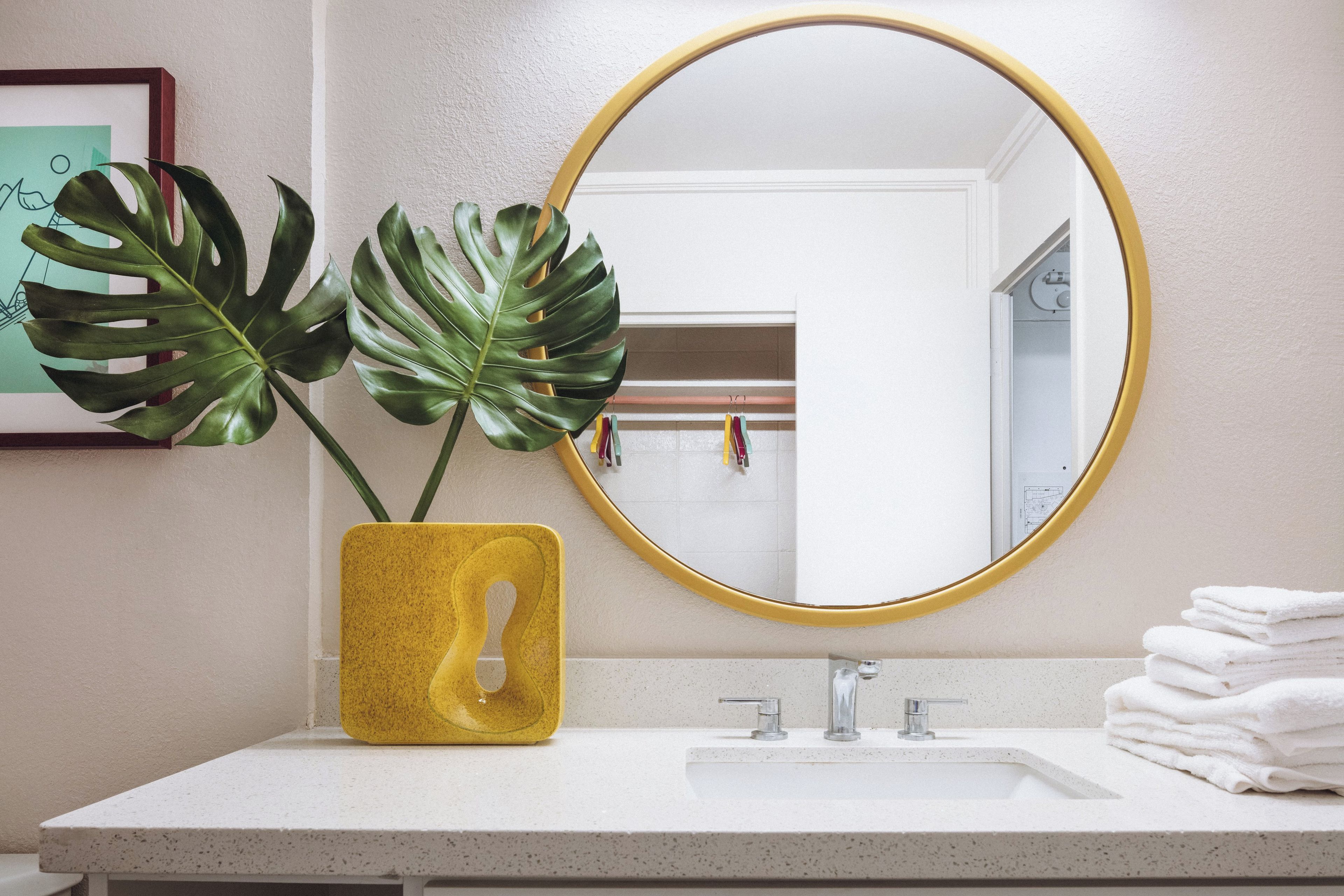 A bathroom with a white sink, a yellow vase with a plant, and a round mirror with a gold frame.