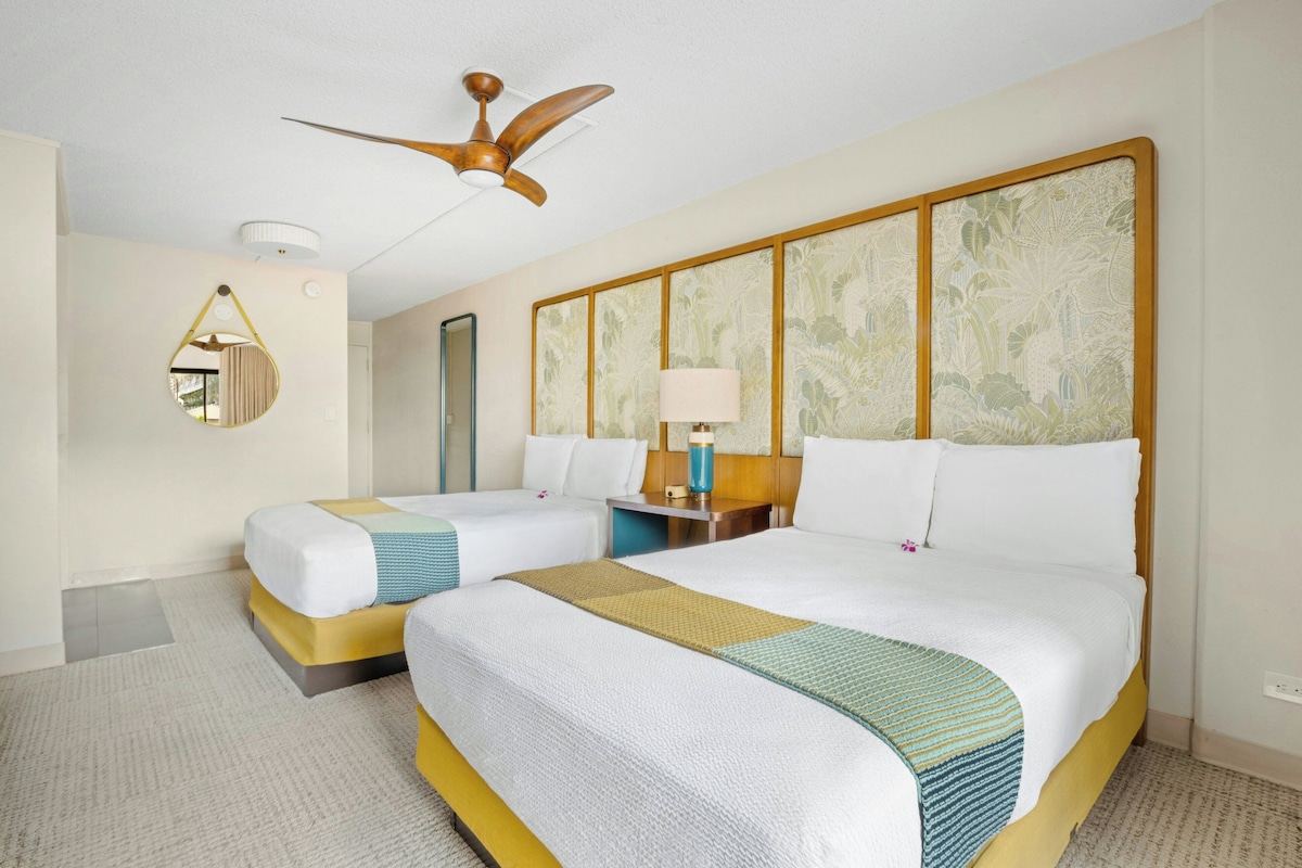 A hotel room with two beds, a ceiling fan, a bedside table with a lamp, a mirror, and a wall with a floral pattern.