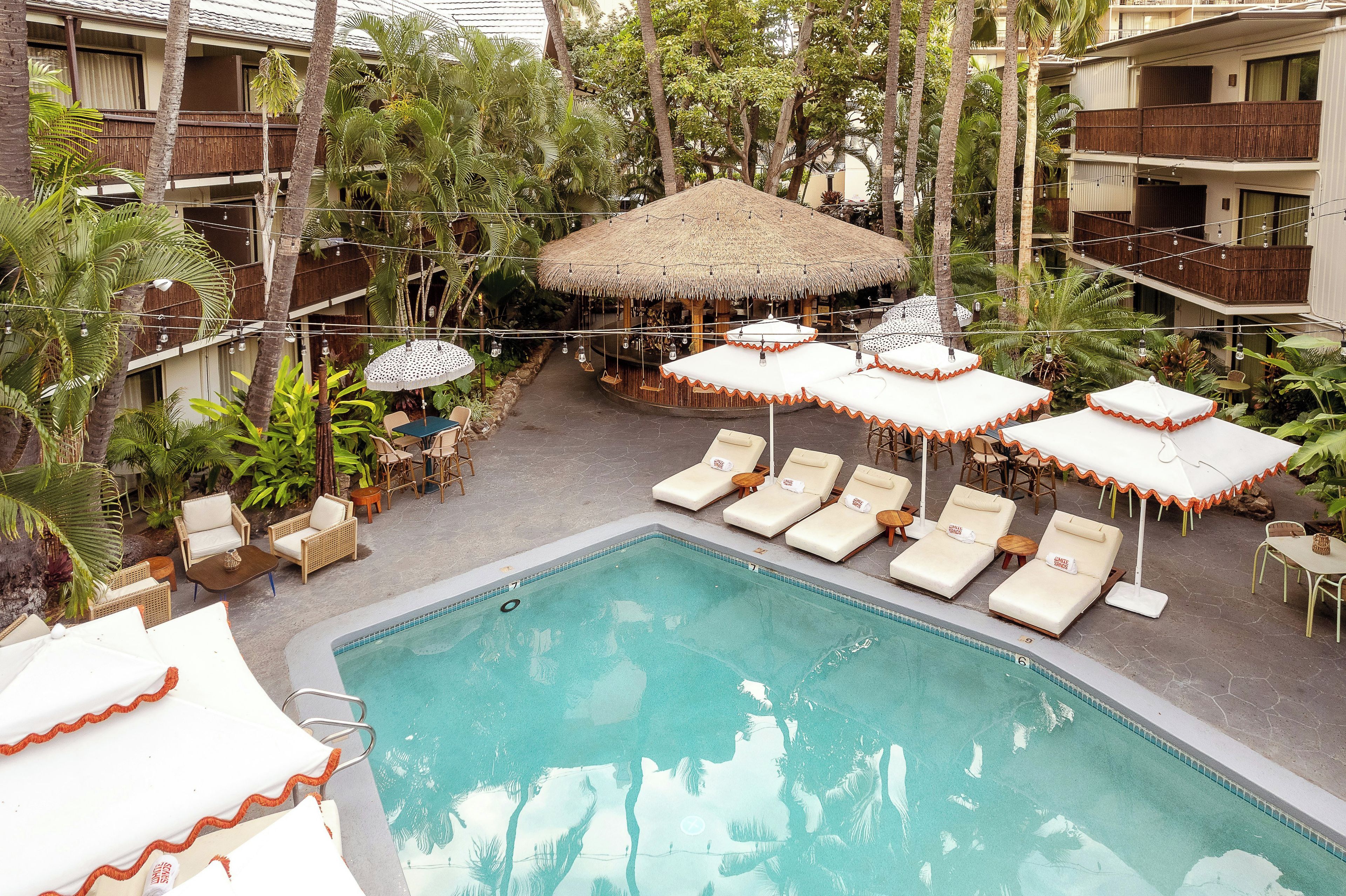 Un espace piscine agrémenté de chaises longues et de parasols, entouré de palmiers et bordé d’un bâtiment doté de balcons.