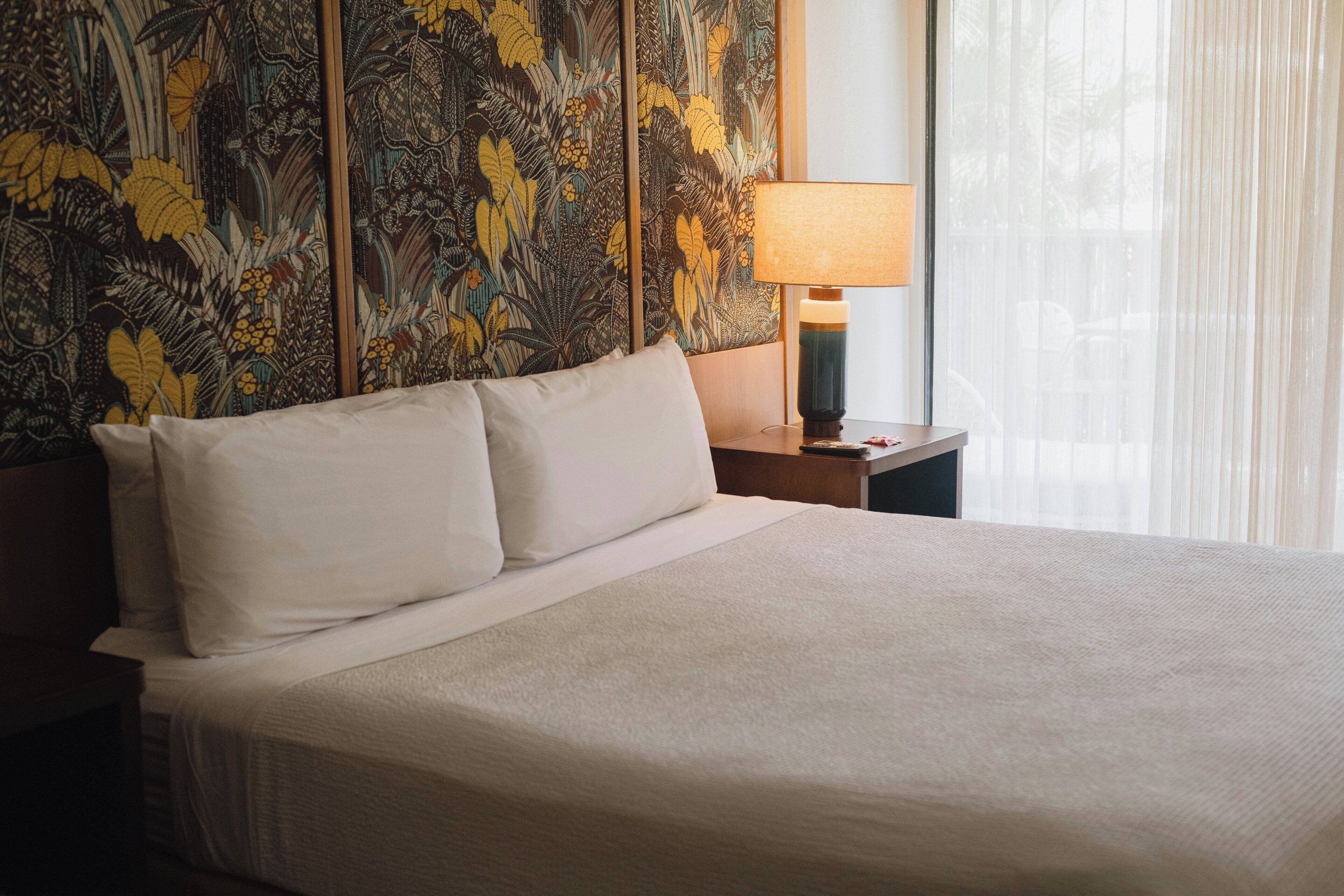 A hotel room with a large bed, a nightstand with a lamp, and a wall with a floral tapestry.