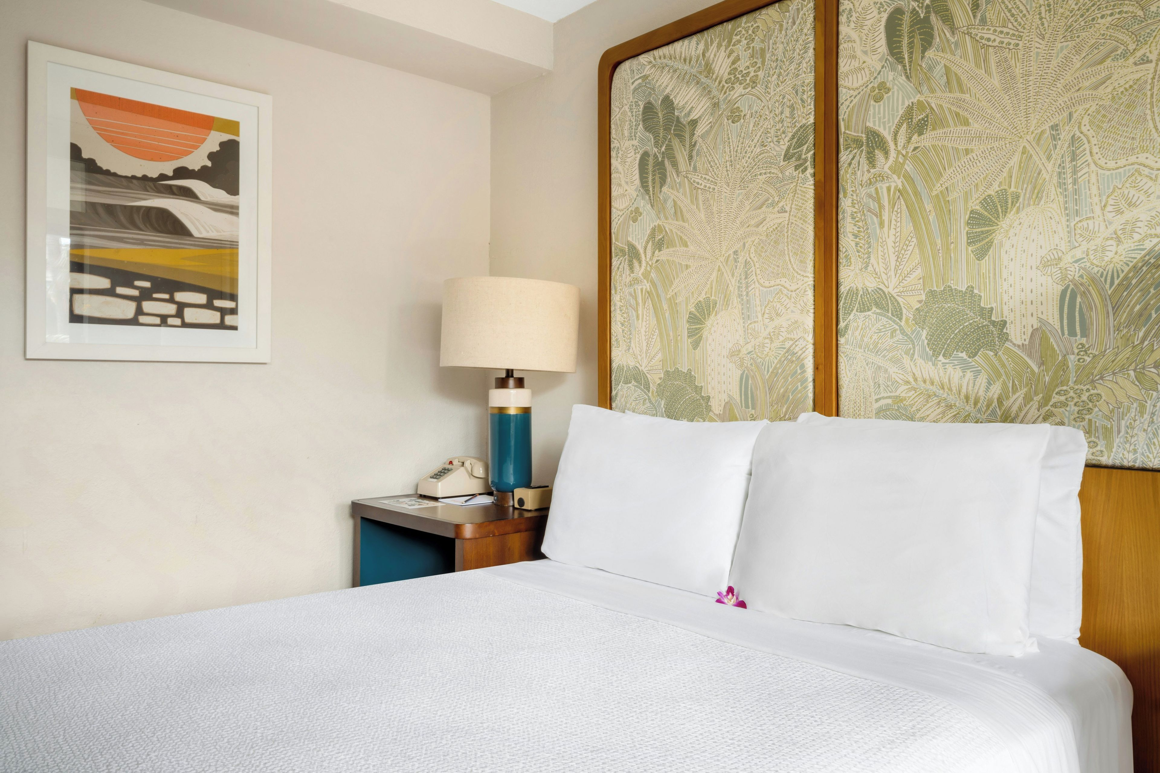 Habitación de hotel con cama, mesita de noche, lámpara, teléfono y un cuadro en la pared.