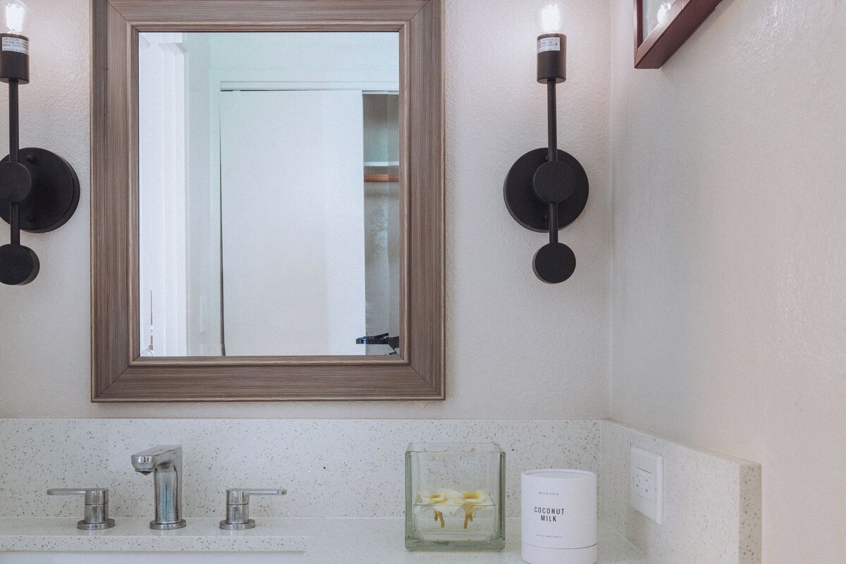 A bathroom with a white countertop, a mirror with a wooden frame, two wall-mounted lights, a glass container with a candle, and a white bottle labeled "Coconut Milk".