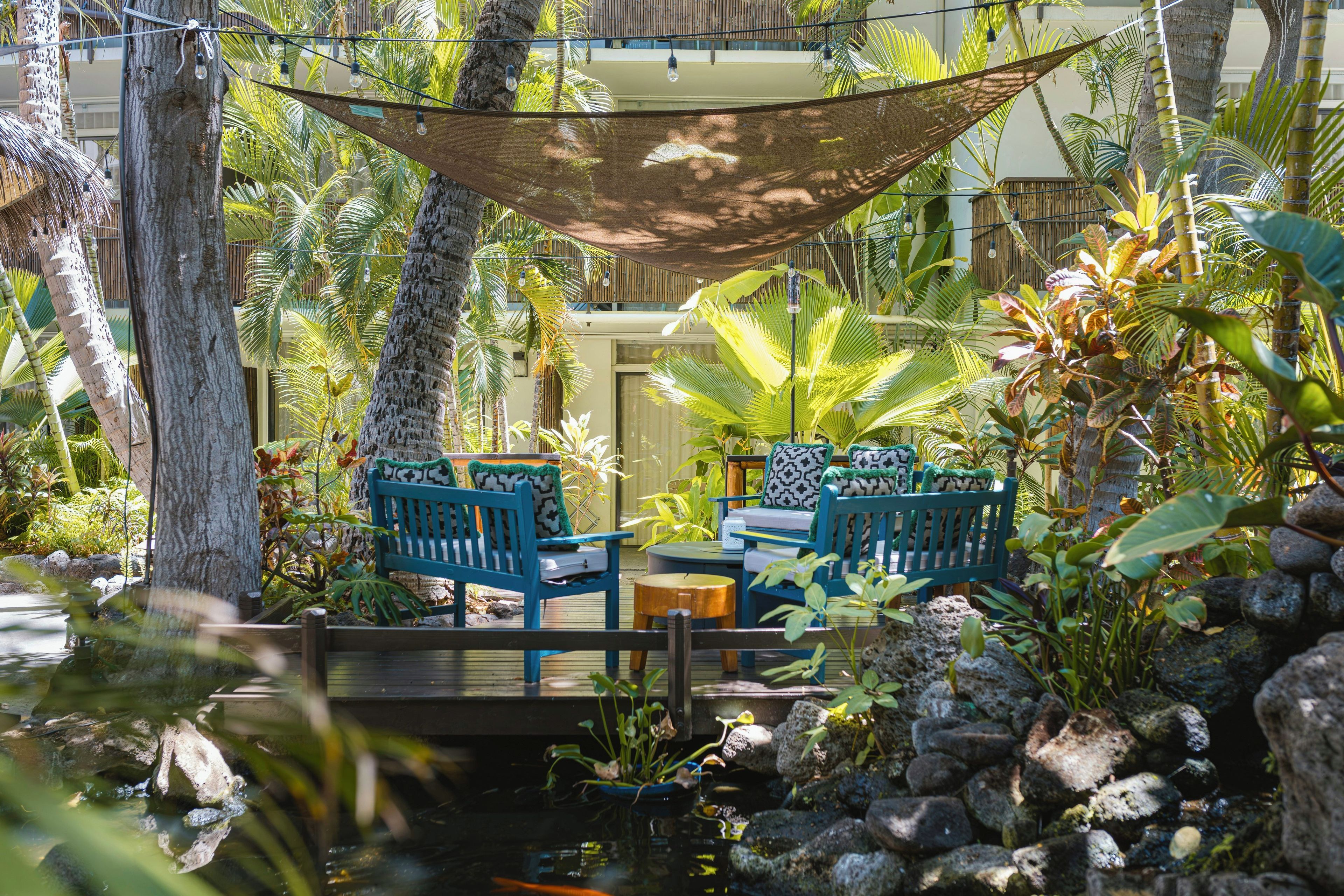 Un espace extérieur paisible, aménagé avec deux chaises bleues et une table, entouré d’une végétation tropicale luxuriante et agrémenté d’un petit élément décoratif aquatique.
